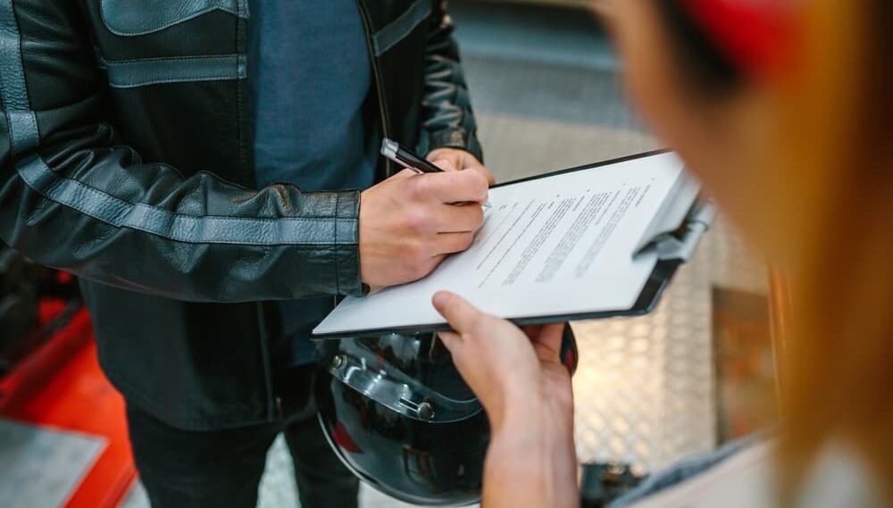 An unrecognizable biker man wearing a leather jacket and holding a helmet, signing an insurance policy to receive his repaired motorcycle.