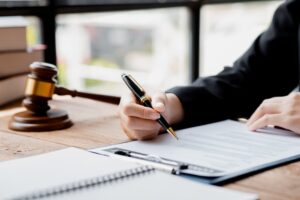 A lawyer sits in his office at a table with a small gavel, symbolic of court proceedings. Justice scales are nearby, and lawyers are drafting a contract for the client. 