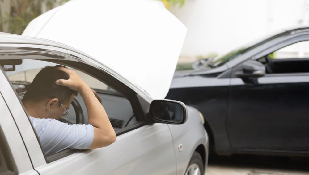 man driver in car accident and holding his head near broken car on the road after a car accident