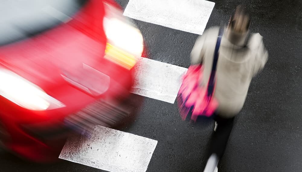 Blurred red car speeding toward a pedestrian with a pink bag crossing a crosswalk, conveying a sense of imminent danger.