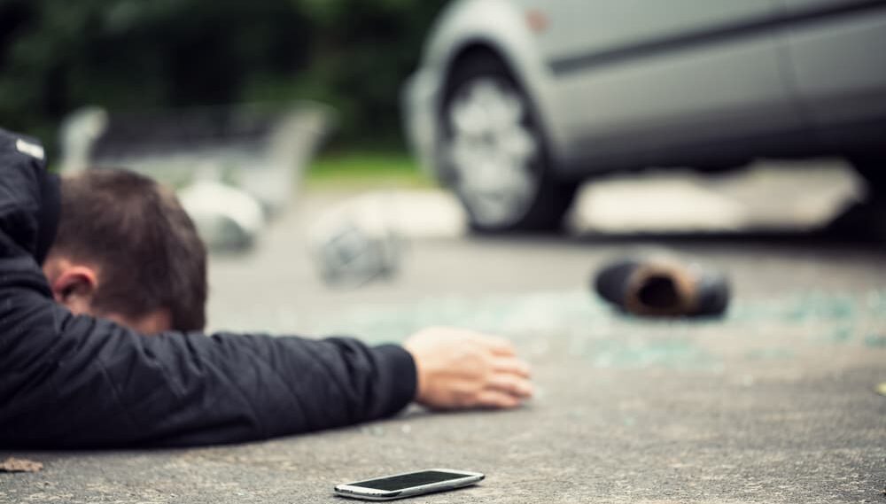 Peatón herido en el suelo tras ser atropellado, con un teléfono caído y un auto cerca.